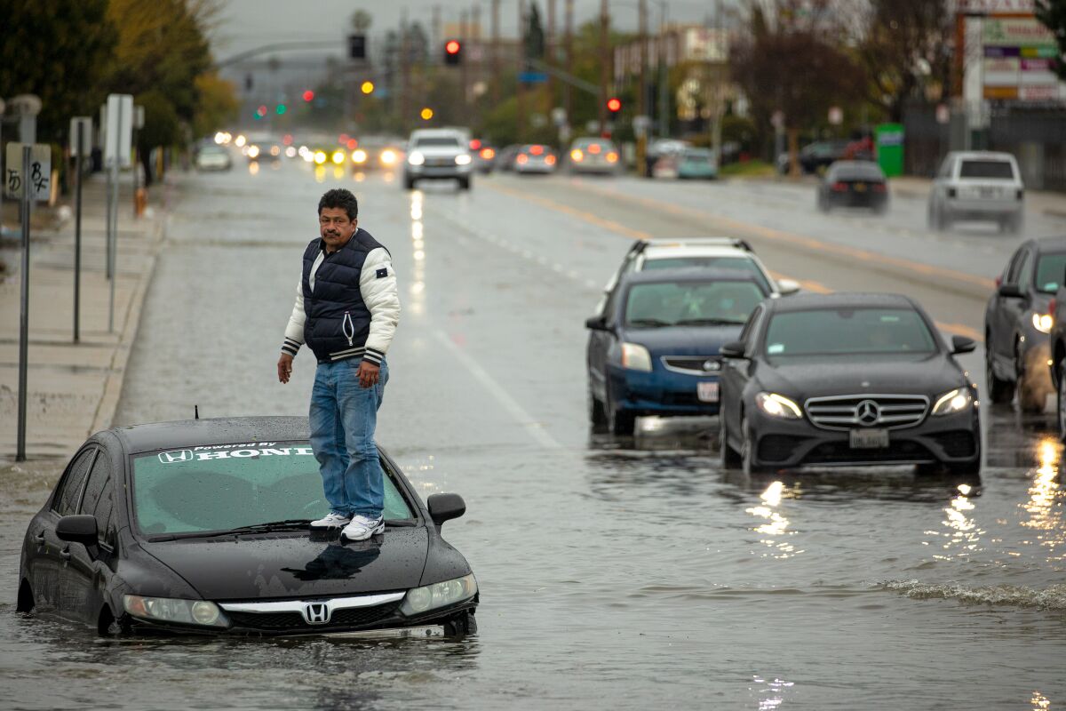 A man stands on his car which is partially submerged in floodwater on a street with several other cars in the background.