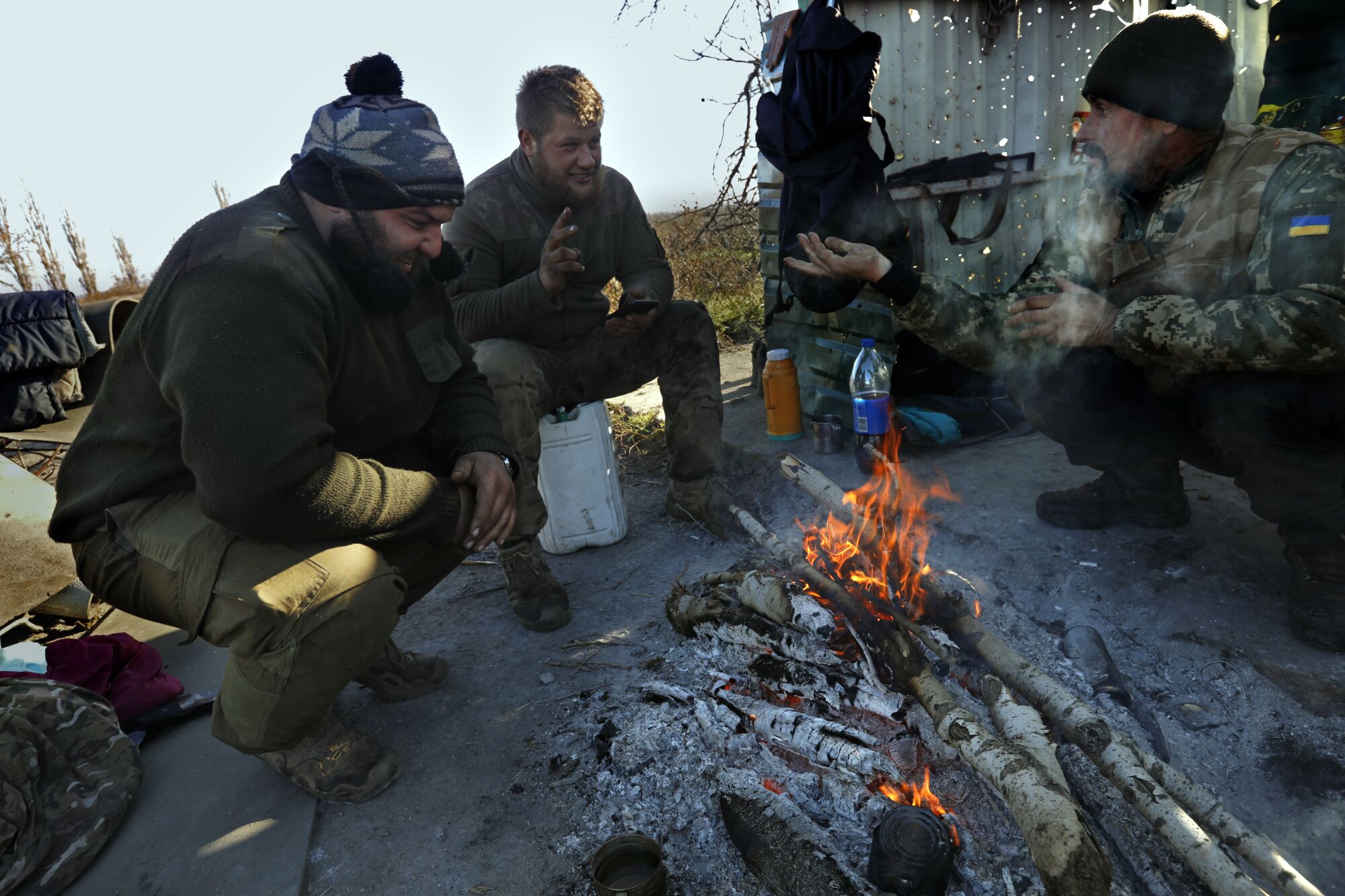 Members of a Ukrainian Army tank platoon get warm by a fire beside a bus stop riddled with bullet holes.