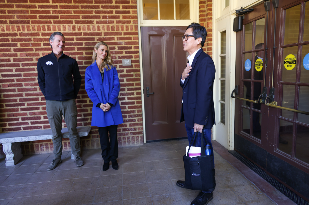 A man and woman stand in front of a brick wall as another man stands near them in front of a door holding a bag.