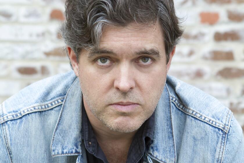 Man with wavy salt-and-pepper hair in blue shirt and denim jacket, seated in front of a brick wall.