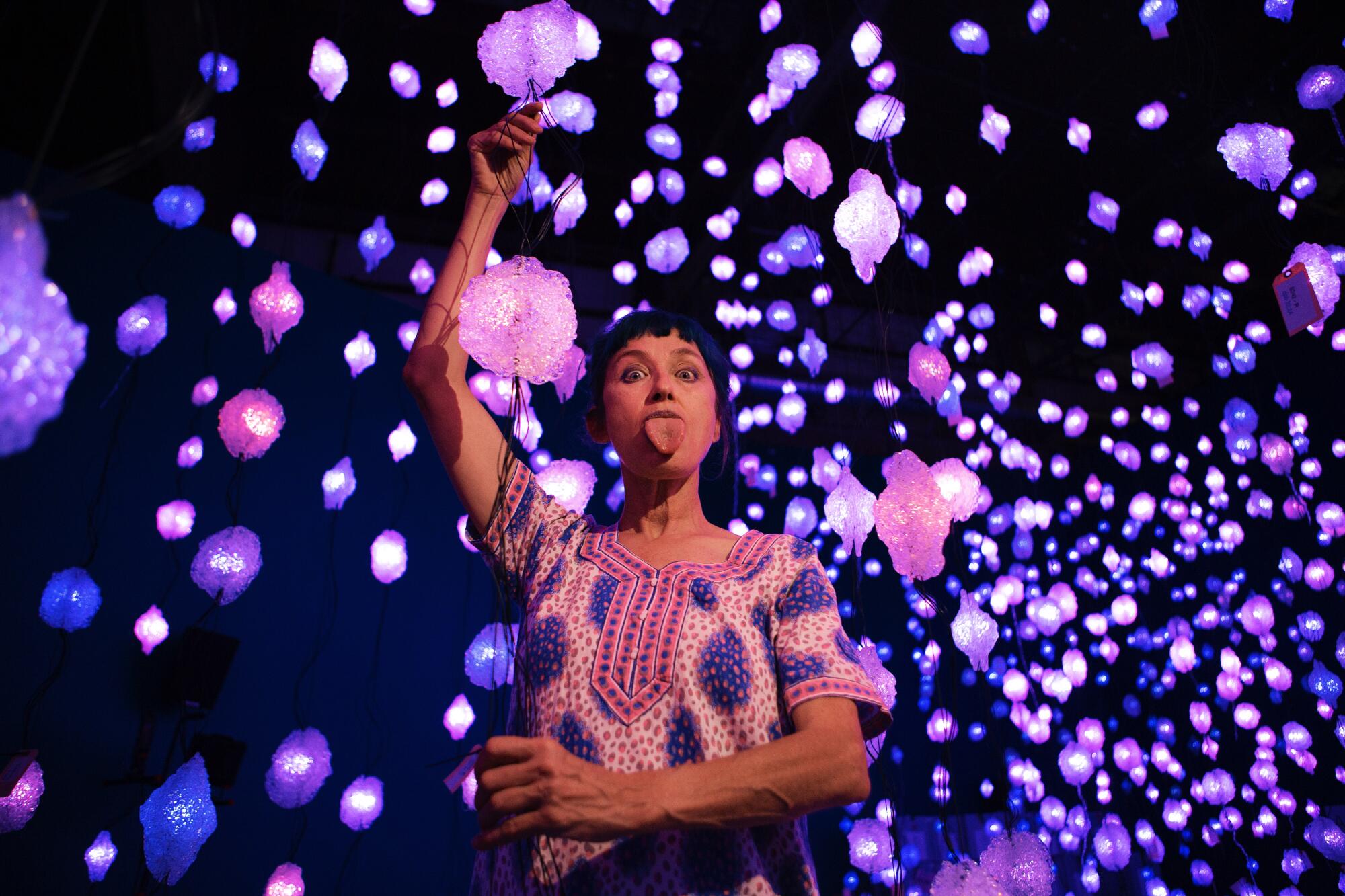 Pipilotti Rist sticks out her tongue while standing amid dozens of pink and purple lights that dangle from the ceiling.