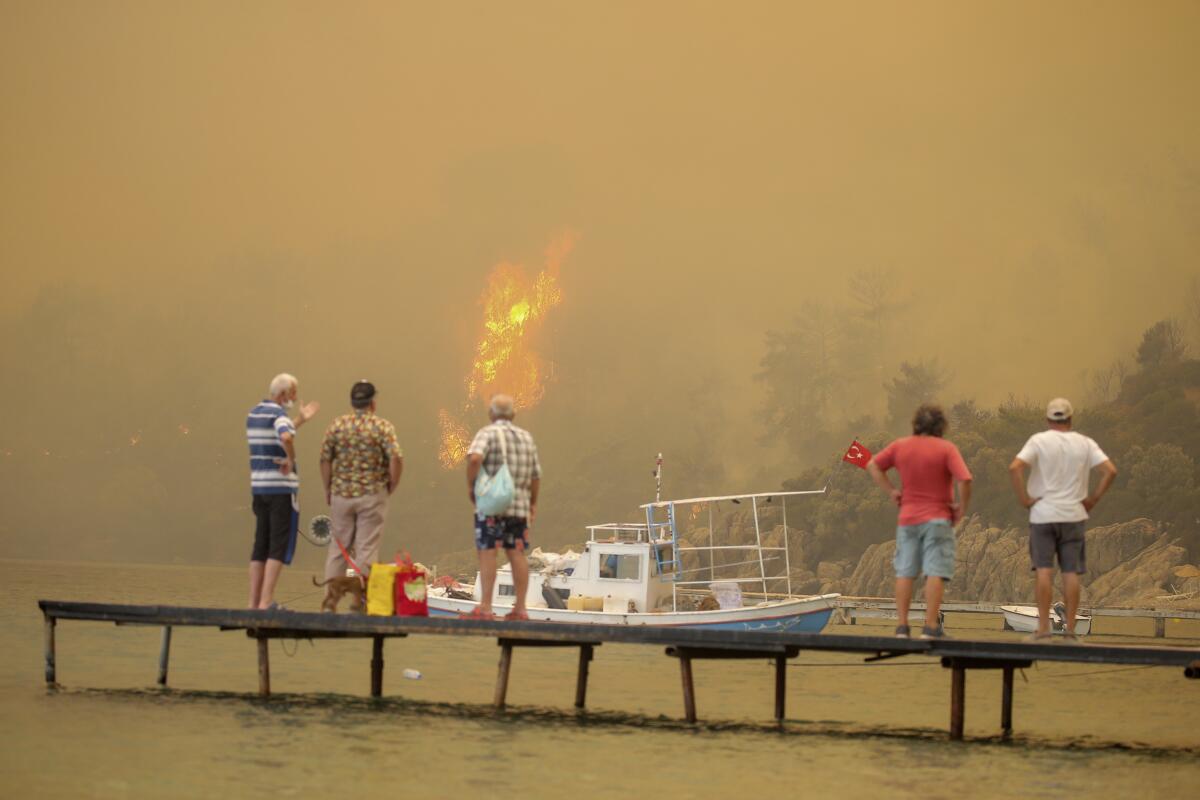 People watch on a dock as a fire burns in the distance