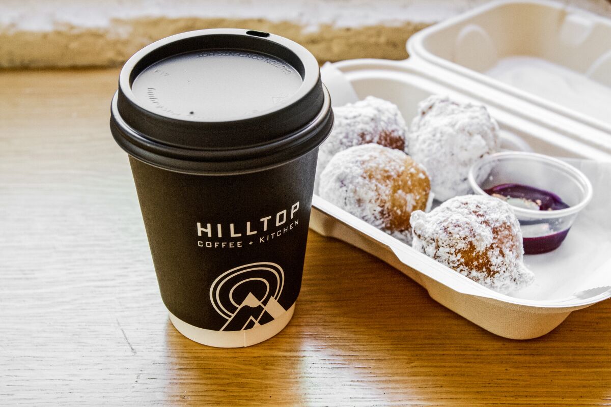Coffee in a paper cup marked Hilltop next to a Styrofoam container of beignets