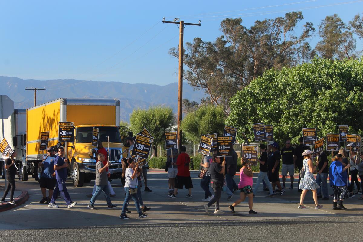 People holding signs walk a picket line on a sidewalk in front of a gate in a fenced-in property as a truck waits to exit