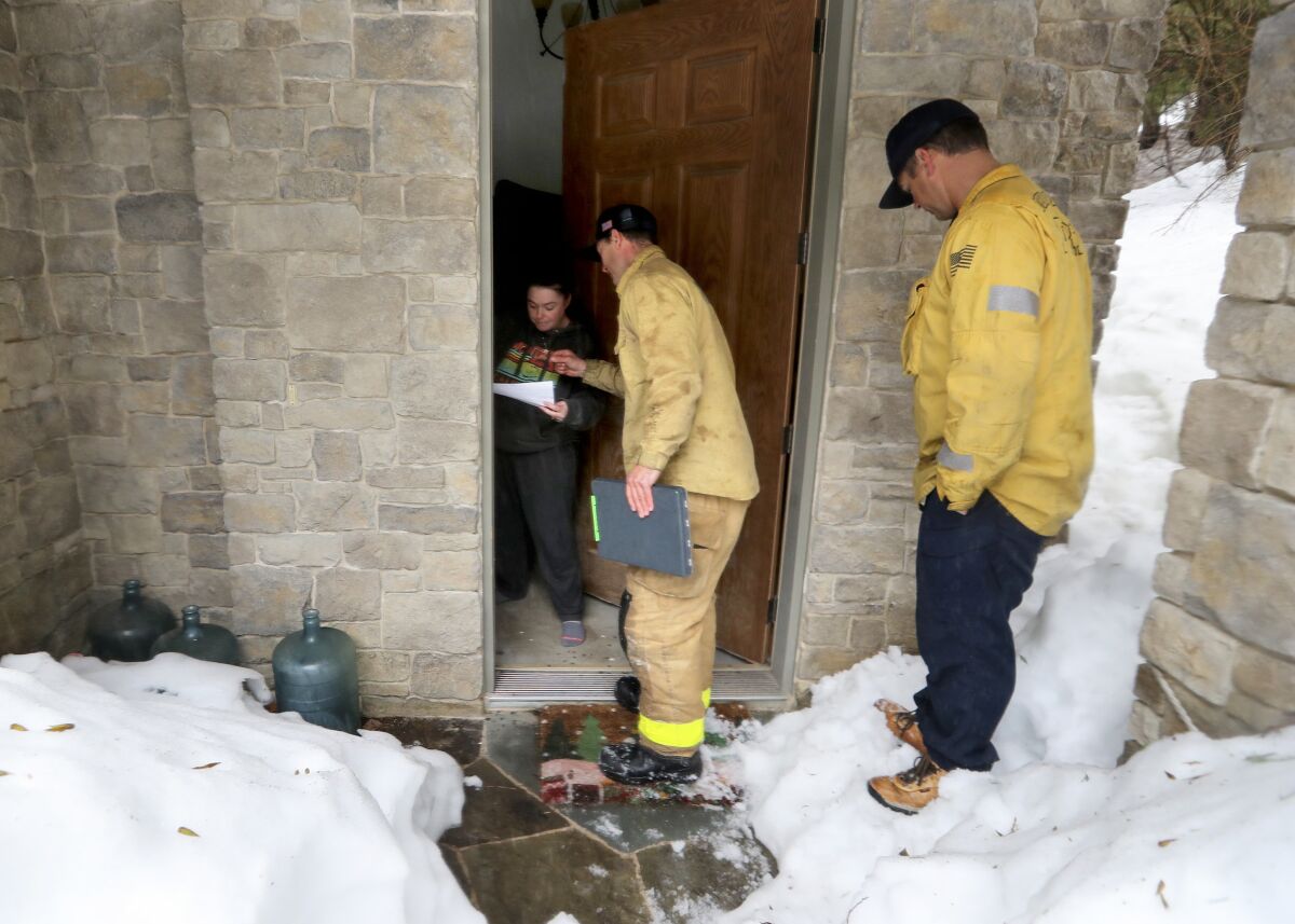 Two firefighters stand outside the door to a stone house where a young woman stands in the door looking at a piece of paper.