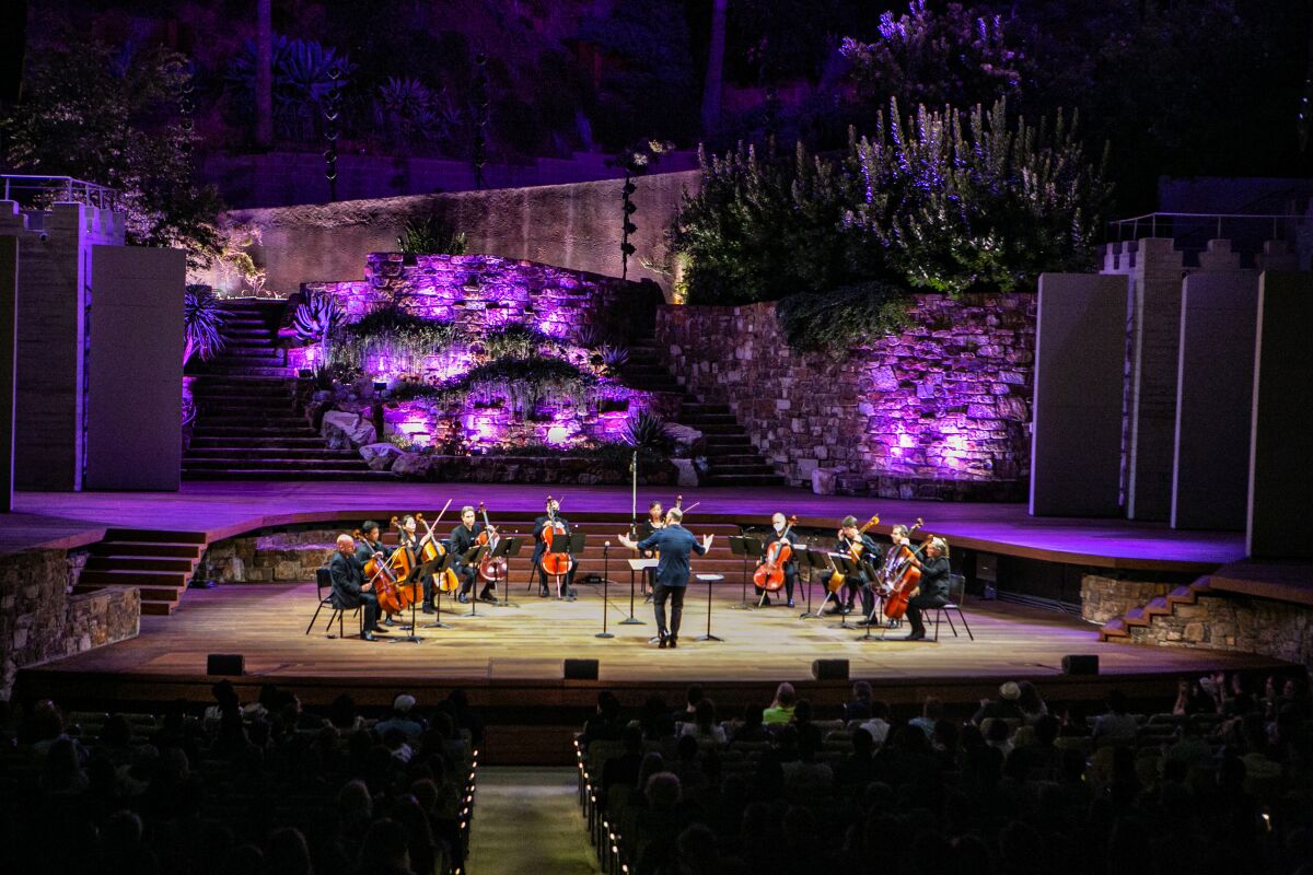 A conductor faces a semicircle of cellists on an outdoor stage.