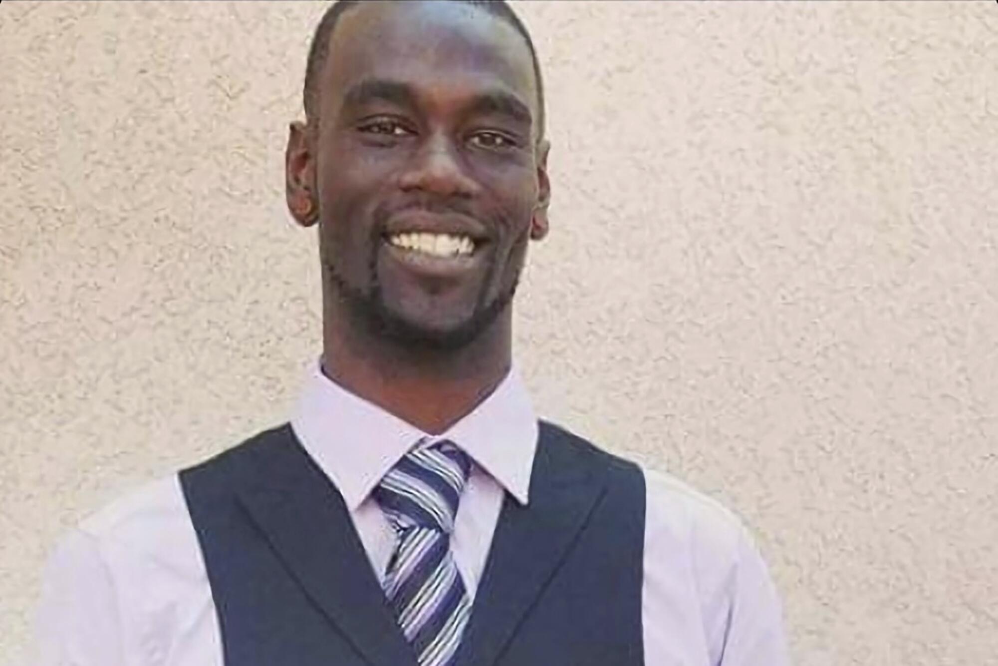 A smiling young Black man, pictured from the shoulders up in a lilac shirt, striped tie and gray vest