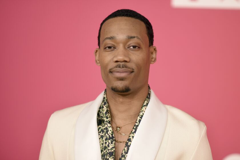 Tyler James Williams poses in a white blazer and patterned shirt against a pink backdrop.