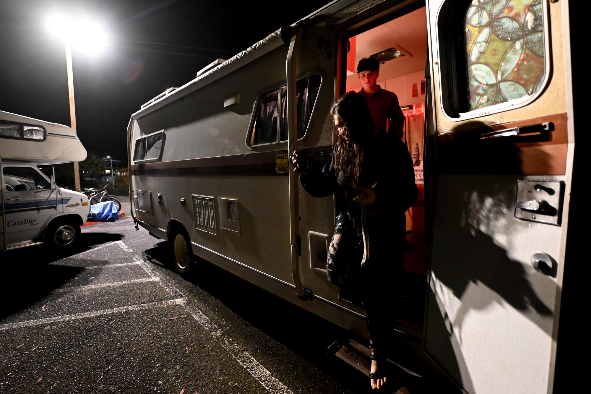 Two people stand in the doorway of a motor home in a parking lot at night.