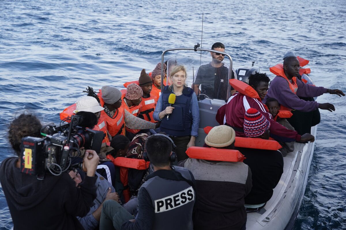 A man points a camera toward a woman holding a microphone on a boat amid a group of seated men