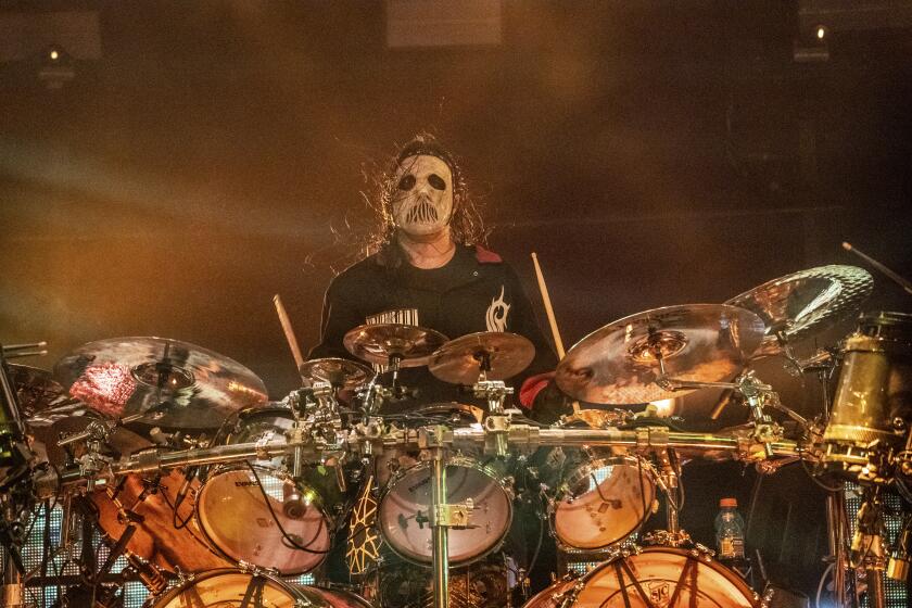 A man with long hair, black and white face paint and a black shirt sits behind a drum kit on a stage