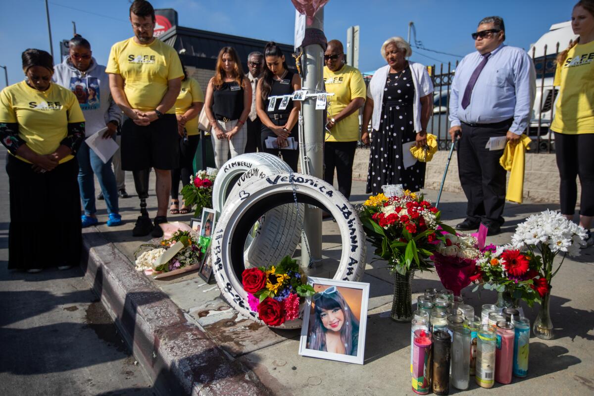 People gather on a sidewalk around a memorial with flowers, candles and portraits.