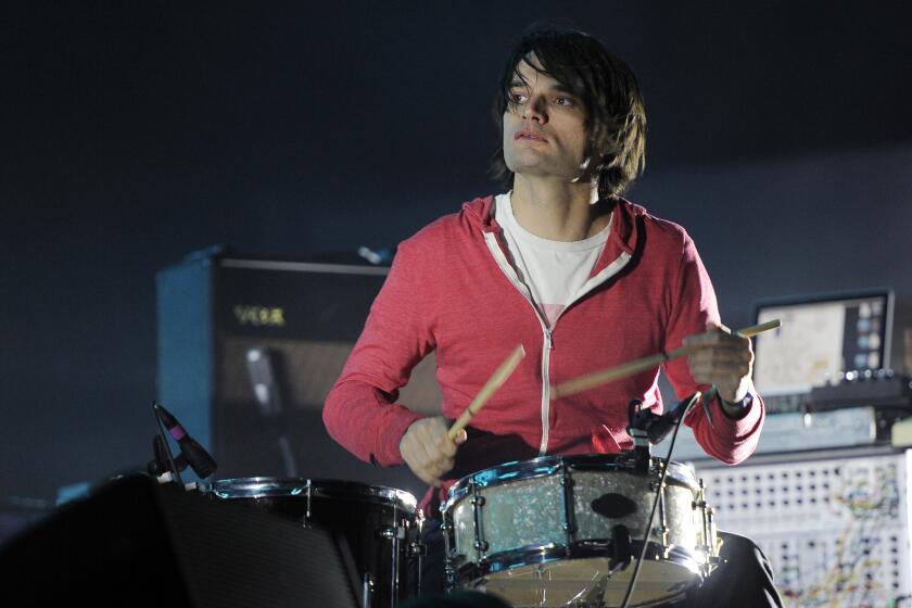 A man with dark hair in a red zip-up hoodie sitting at a drum kit playing a drum