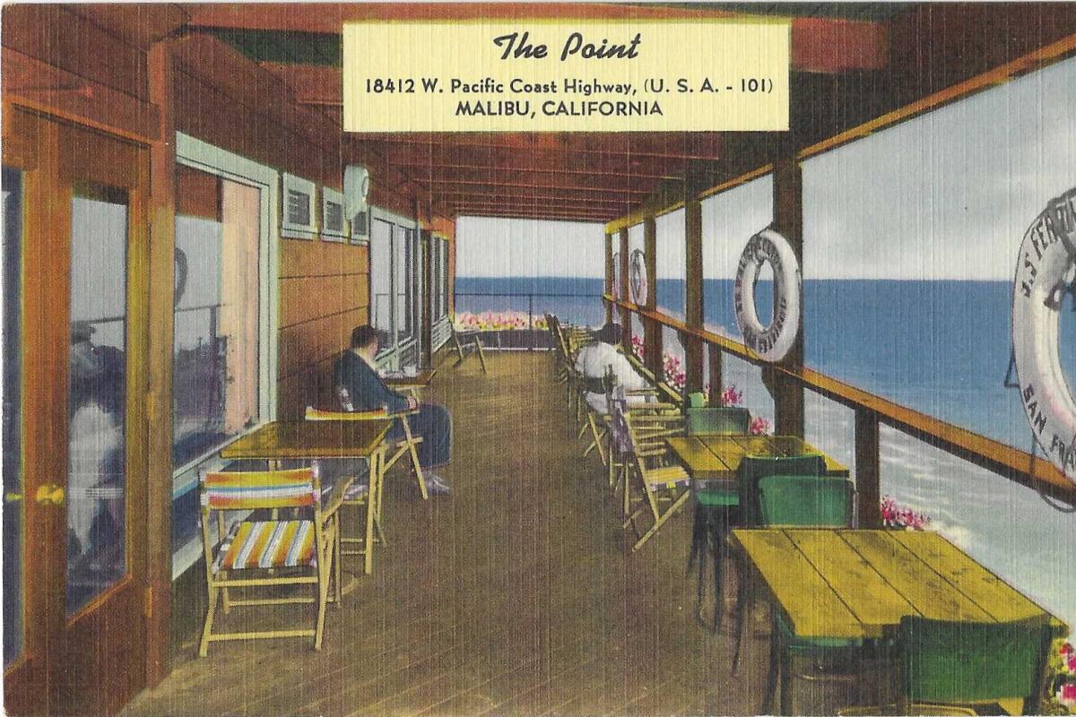 People sit at cafe tables on a deck overlooking the water