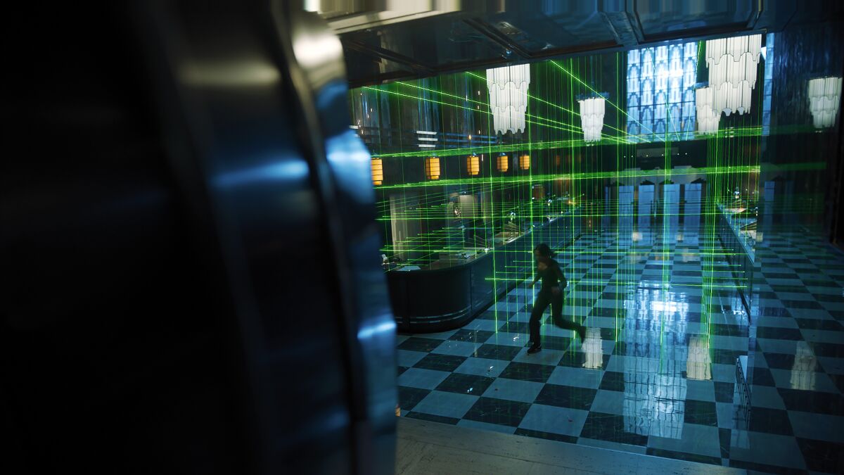 A woman stealthily walks on marble checkered floors in a high-ceilinged room filled with green lasers.