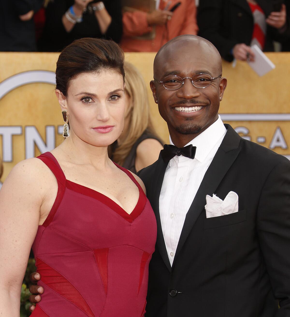 Idina Menzel wears a red dress and Taye Diggs wears a black tuxedo as the two pose for photos at a red carpet event.