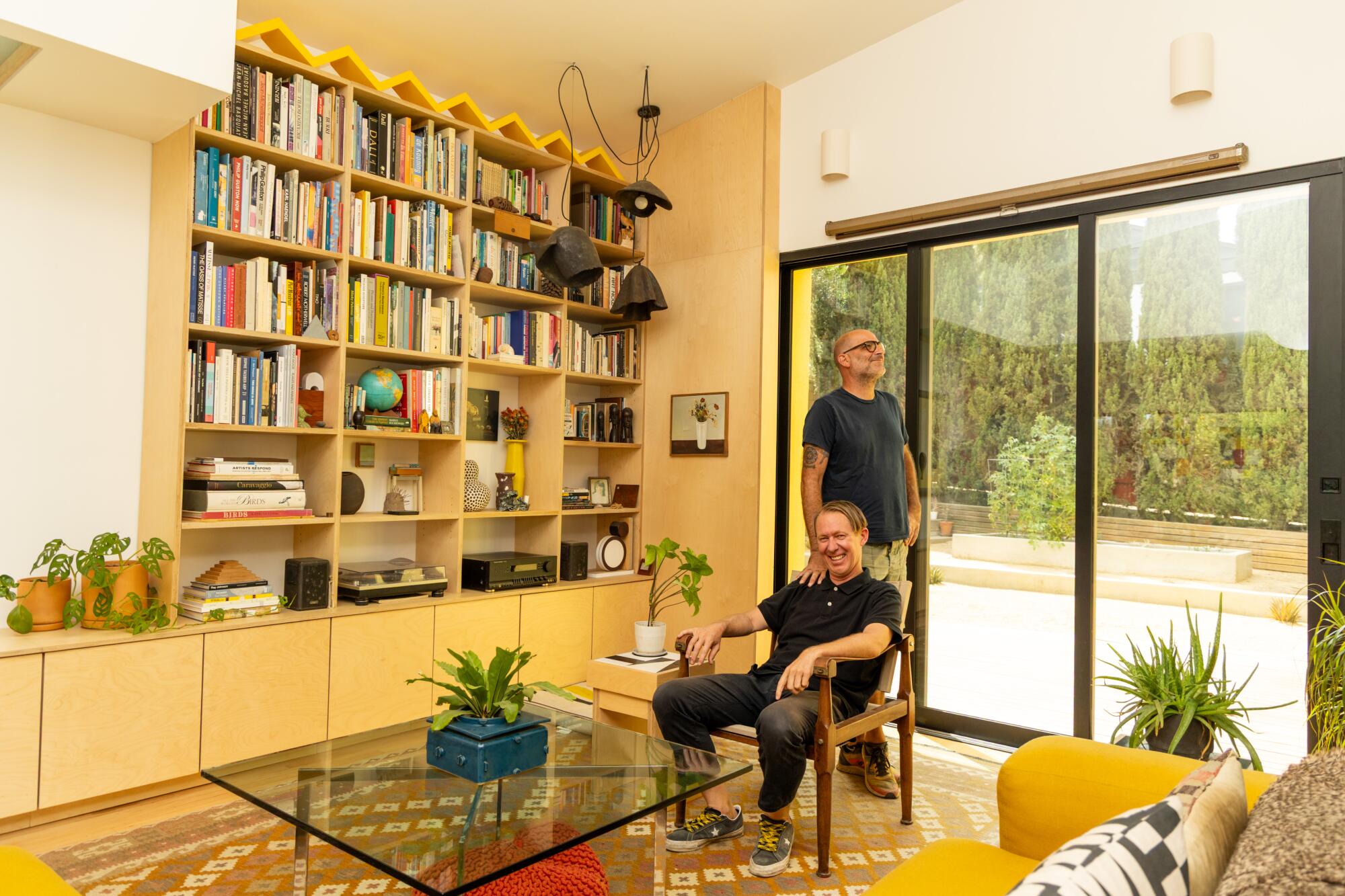 Two people, one seated and the other standing, in a living room space with bookshelves and drawers.