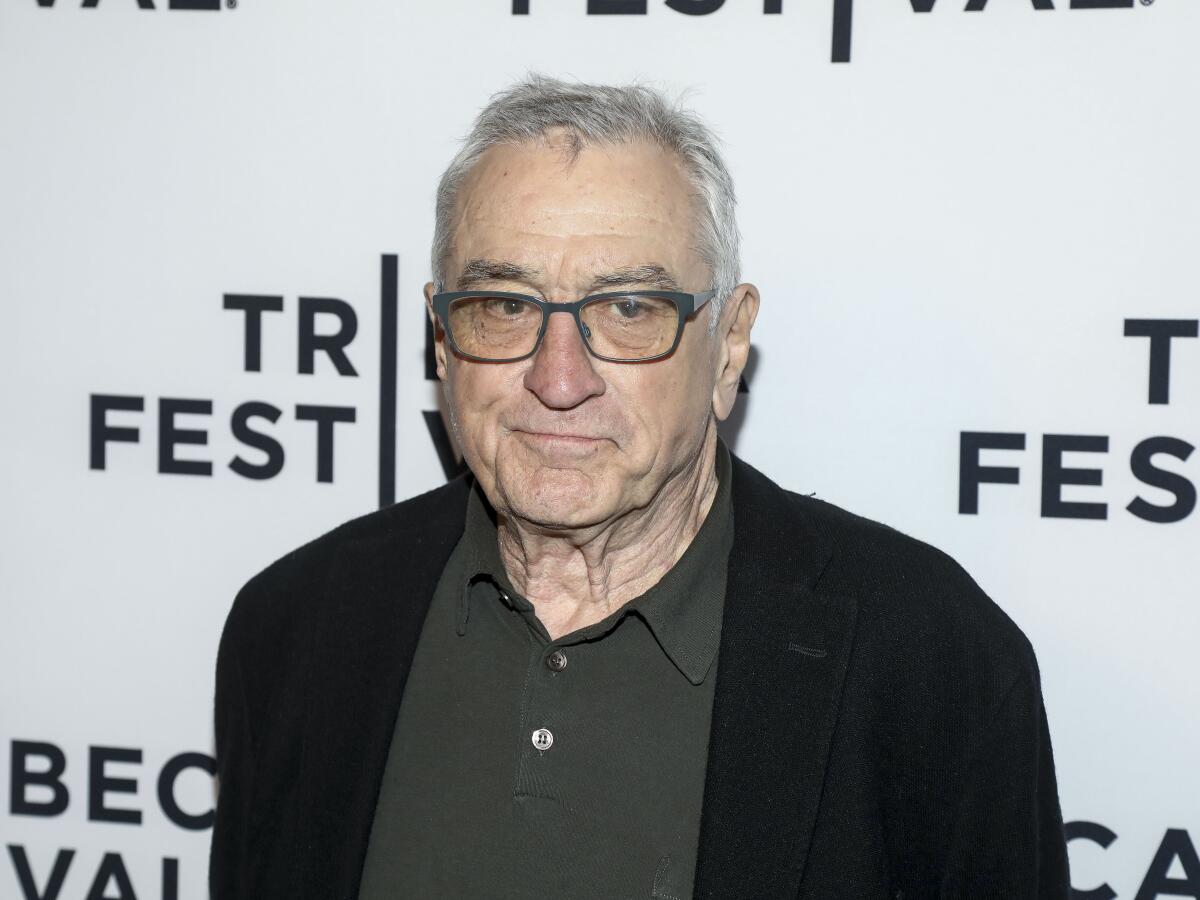 Robert De Niro holds his lips tightly together while posing in a black jacket, dark polo shirt and glasses