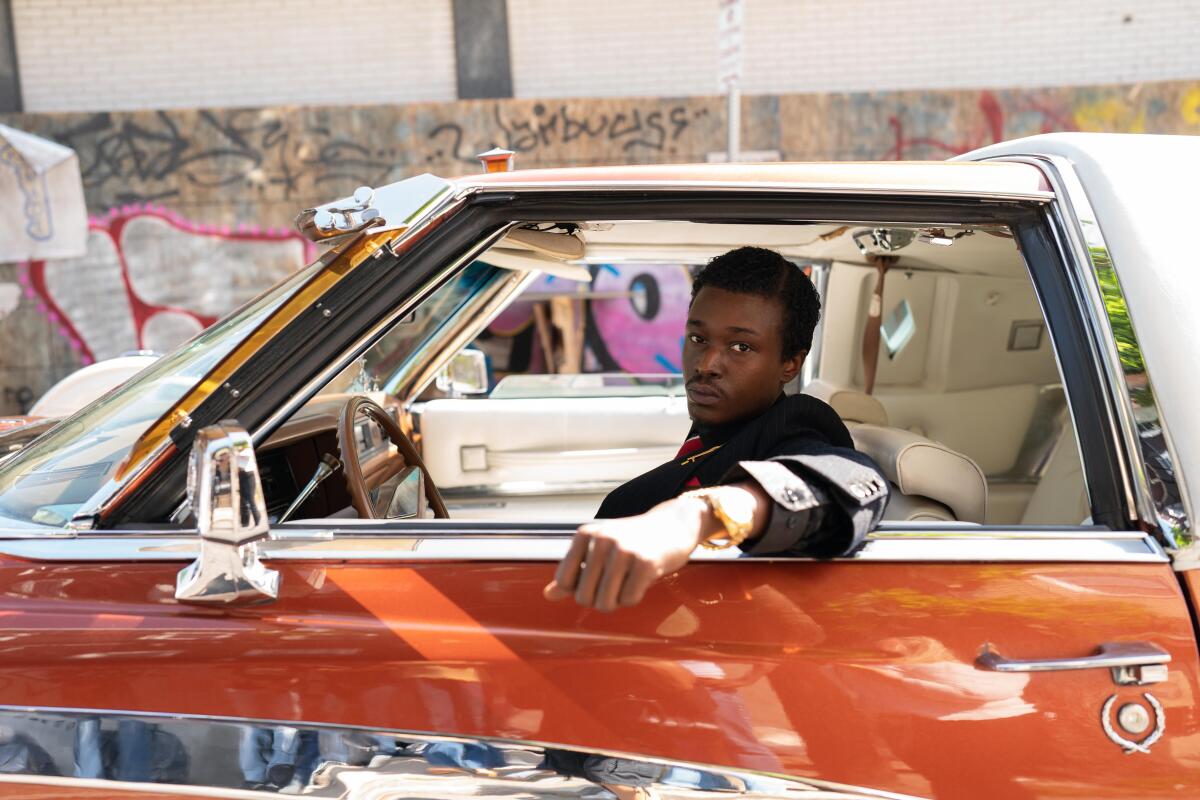 A young man looks out the driver's side window of a burnt orange sports car, directly at the camera