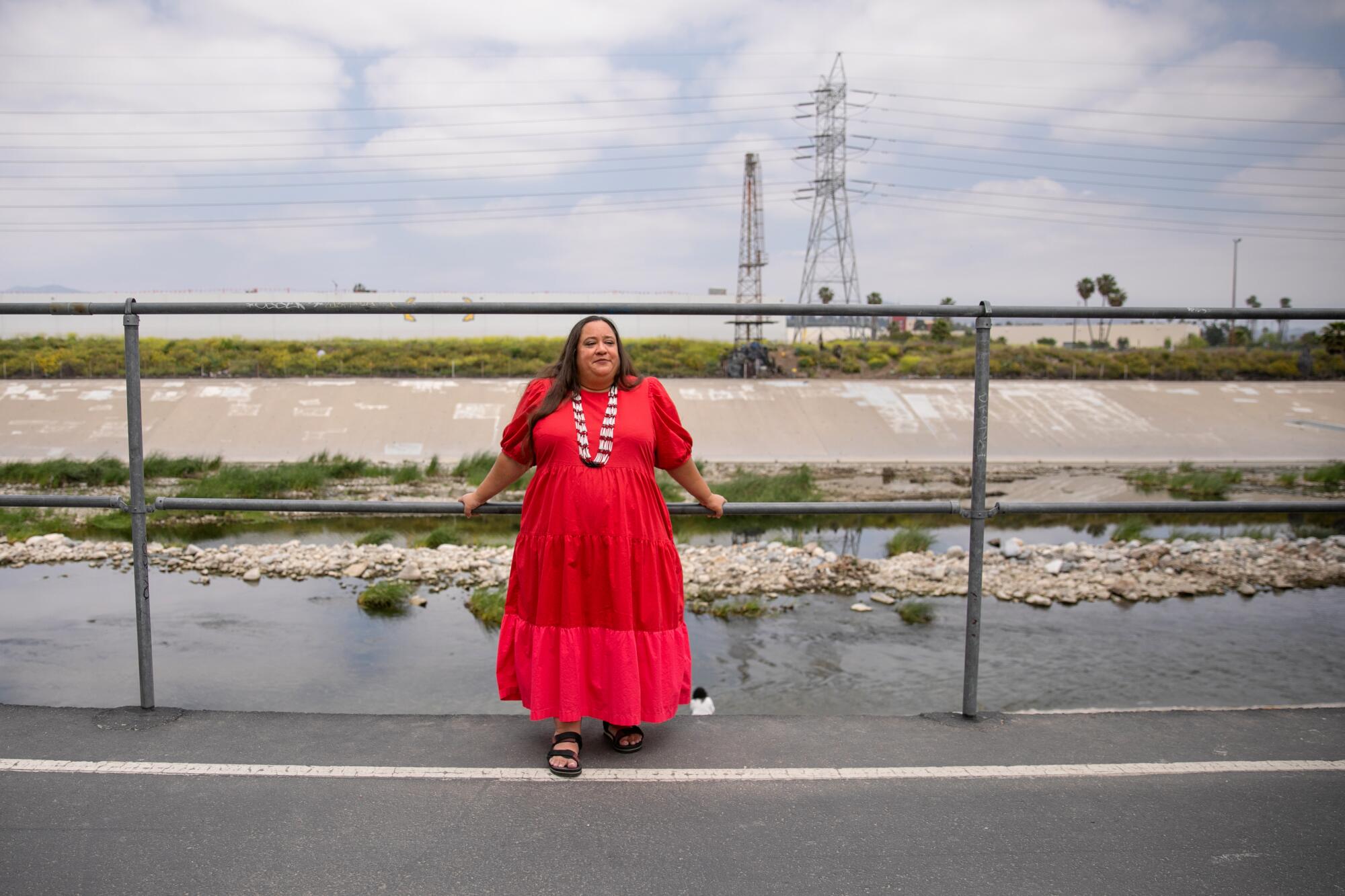 A woman in a long red dress with long brown hair leans against a metal barrier with a concrete-bed river behind it.