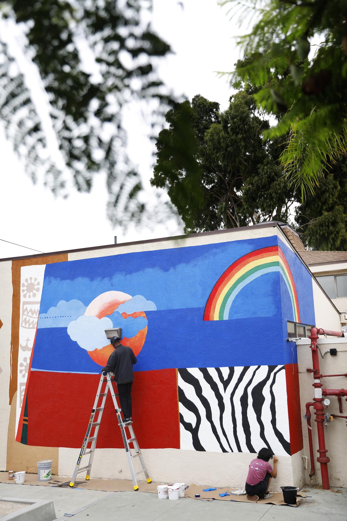 A person on a ladder and one kneeling work on a bright, bold wall mural.
