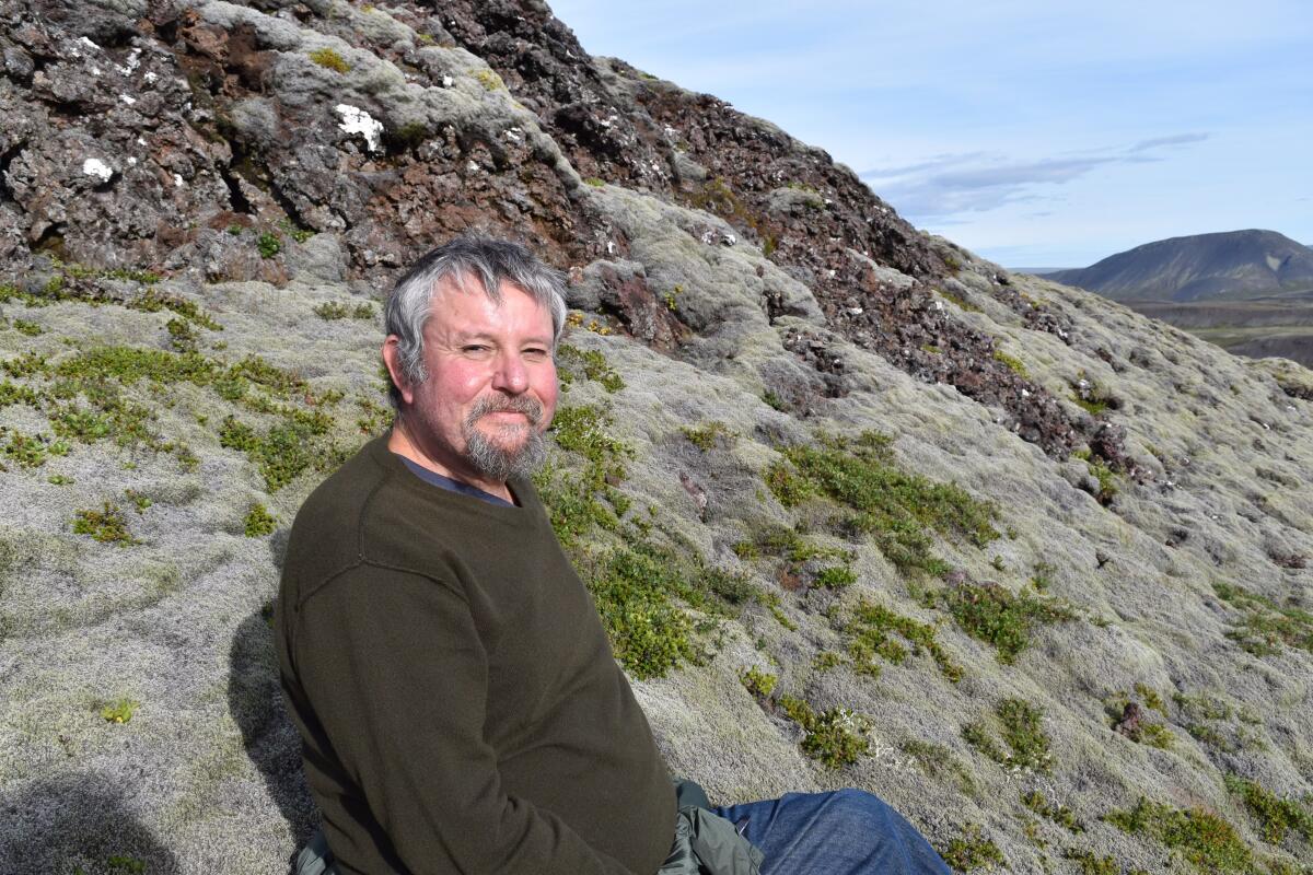 A gray-haired, bearded man sits smiling on a mountainside.