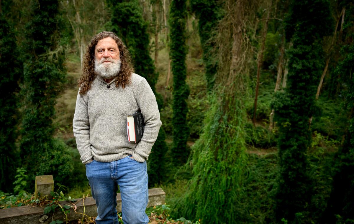 A man with curly brown hair and gray beard stands amid trees, holding a book under one arm