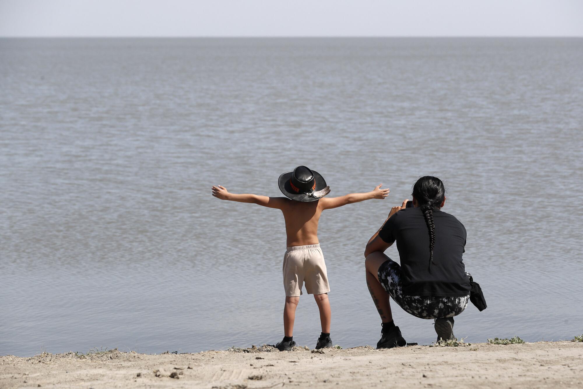 A young boy raises his arms beside a lake as his father kneels beside him.