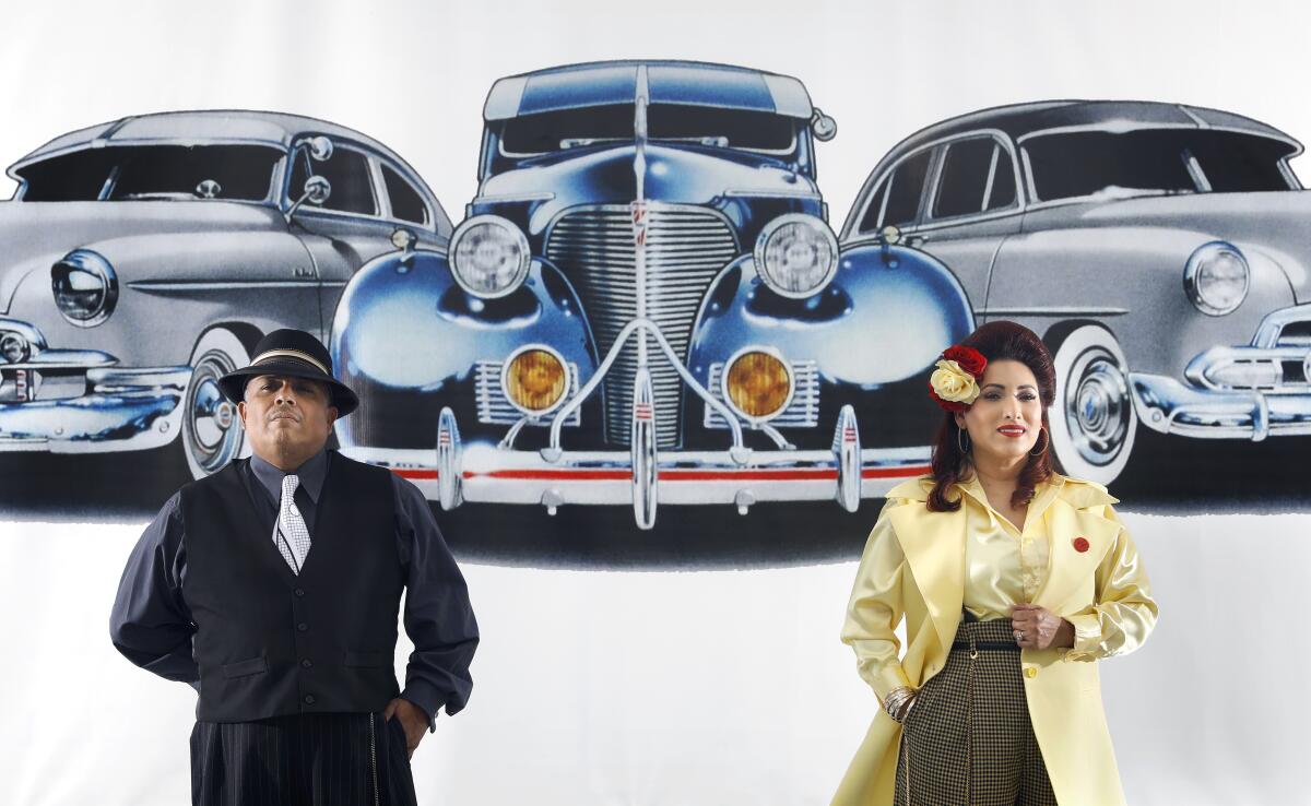 A man and a woman in zoot suit attire — he in black, she in bright yellow — stand before a graphic of classic cars.