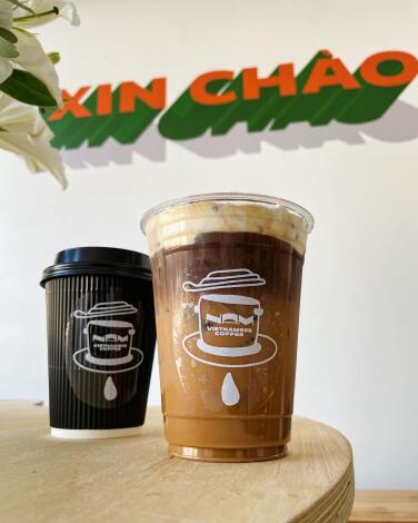 Two Vietnamese coffee drinks, one iced and one hot, in front of a mural that says "XIN CHAO" at Nam Coffee in Hollywood.