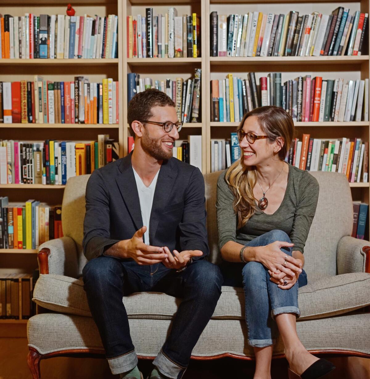A man with a trimmed beard and a woman with long blond hair look at each other against a backdrop of bookshelves.