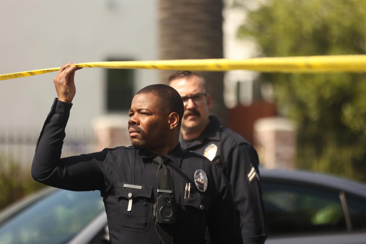 A police officer lifts yellow crime scene tape above his head as he looks beyond it, another officer behind him
