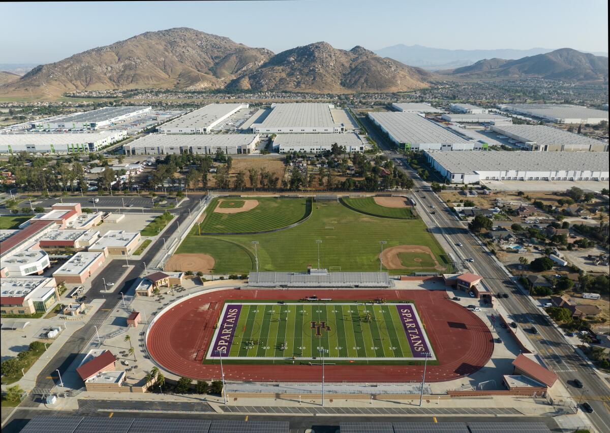 High school athletic fields are seen in the foreground while large warehouses and mountains can be seen in the background.