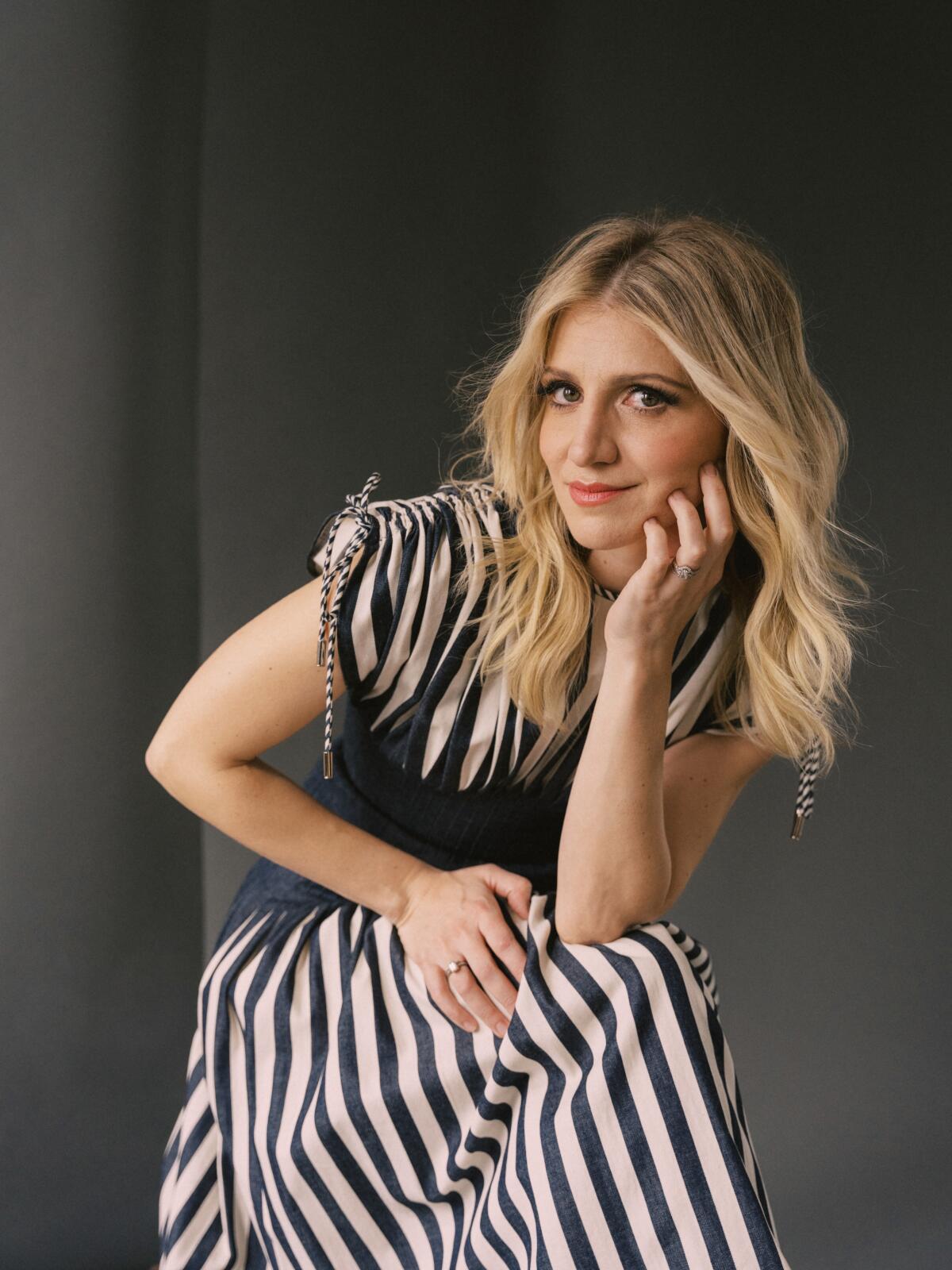 Annaleigh Ashford, in a blue and white striped dress, looks at the camera with her fingers on her cheek.