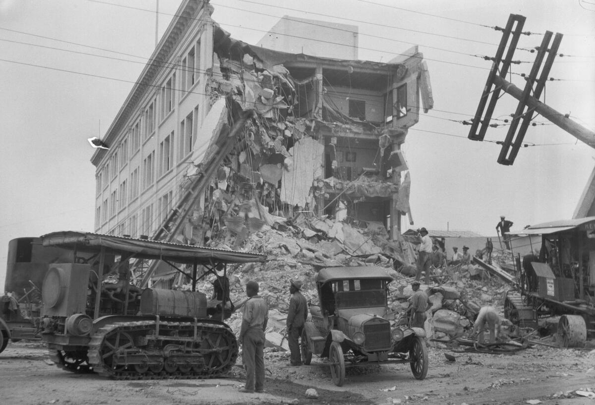 One end of a multistory building is in rubble. A Model T and a type of tractor from the same era are nearby.