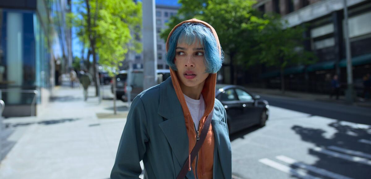 A young woman with blue hair stands on a city street, looking to one side.