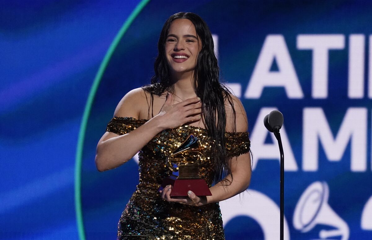 A woman with long brown hair wearing a gold dress, smiling and holding an award