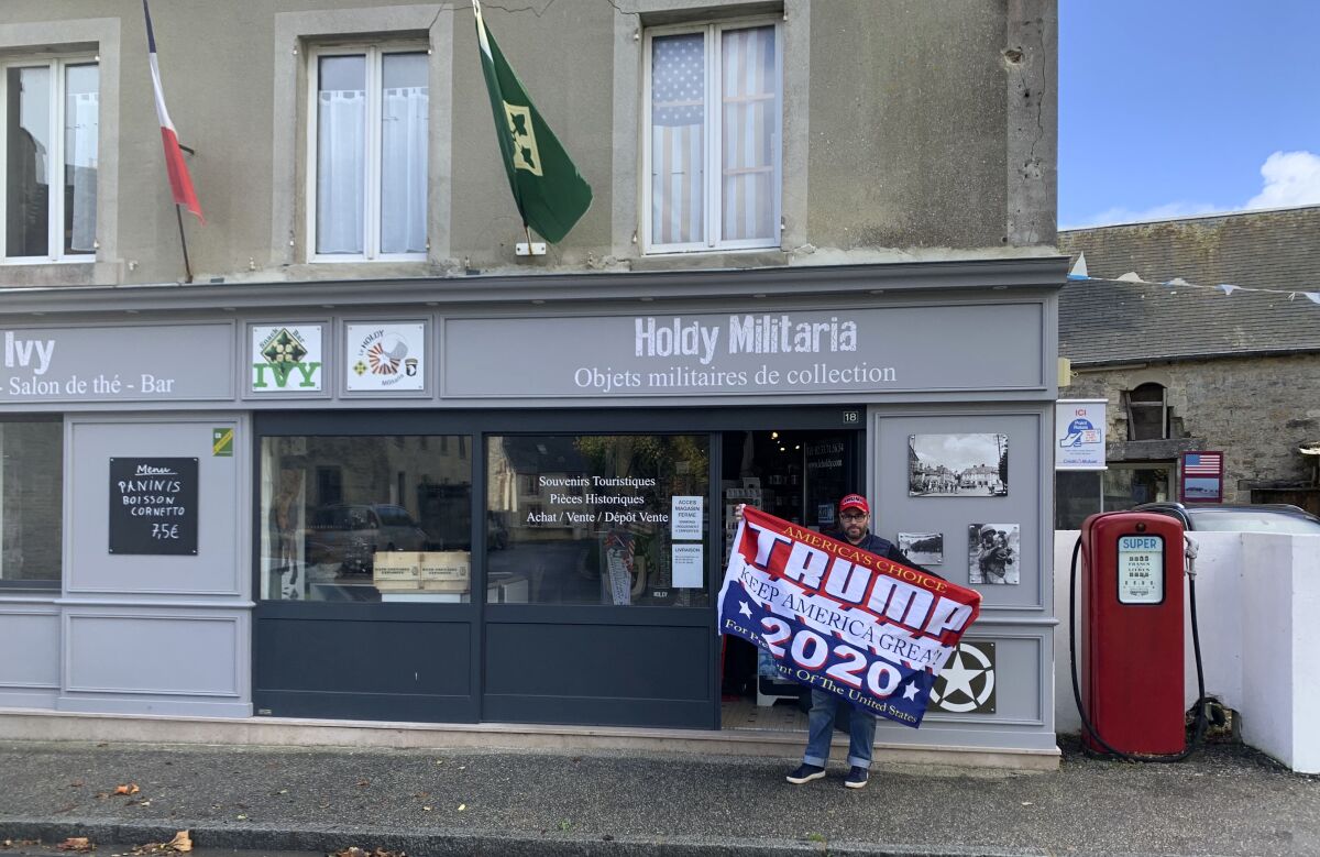Philippe Tanne, of France, holds a Trump 2020 flag outside the military memorabilia store he runs in the Normandy town of Sainte-Marie-du-Mont, one of the sites of D-Day invasion in 1944, Tuesday, Nov. 3, 2020. Tanne, a French former soldier who hopes Trump wins reelection, is among the multitudes of people across the globe for whom the U.S. election is not a far-away happening in a far-away land but an impossible-to-ignore big deal for the planet. (Courtesy of Philippe Tanne via AP)