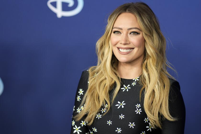Hilary Duff in a black dress with a white flower pattern smiling and posing against a dark blue background