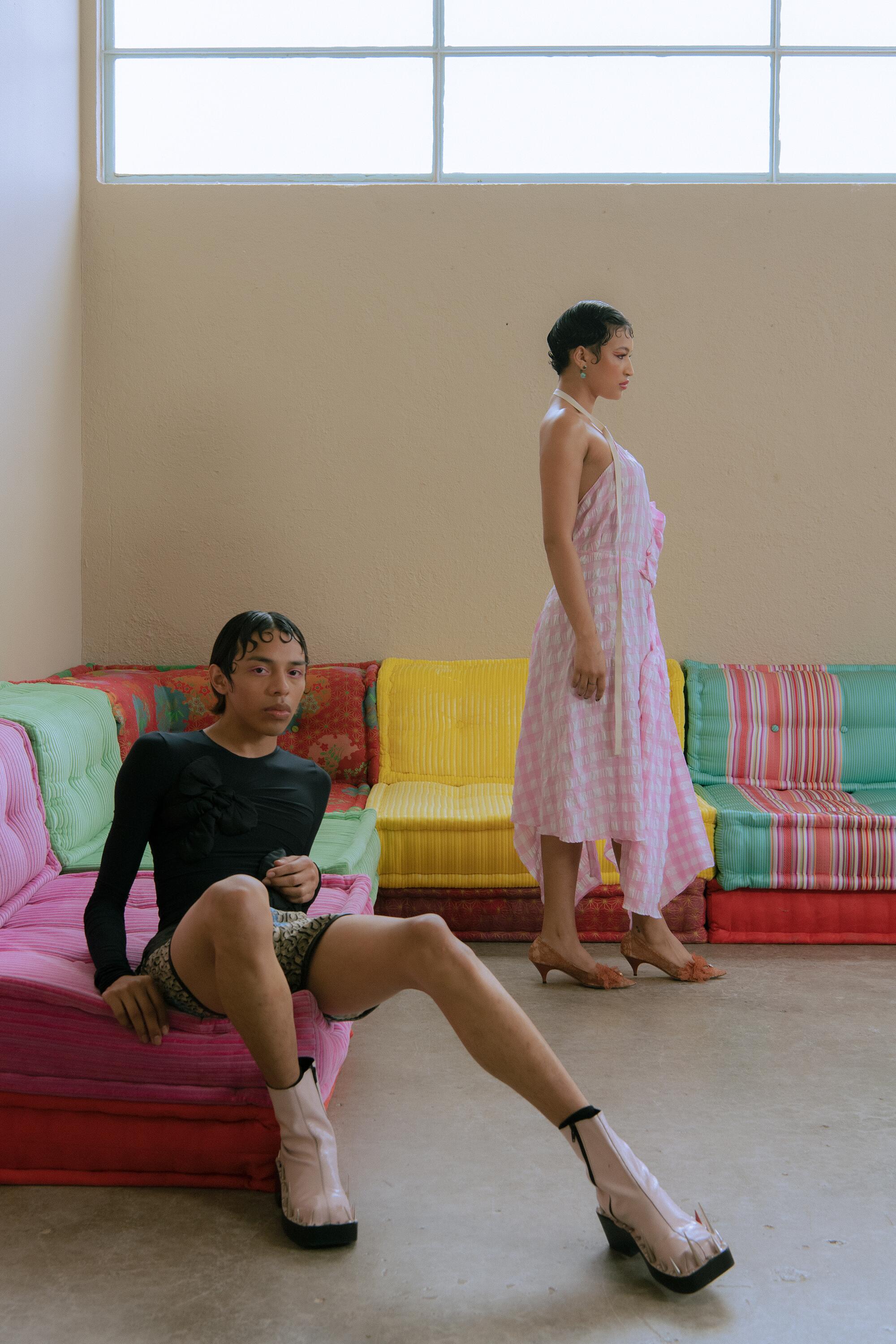 One model sits on a multi-colored, V-shaped couch, while another model stands in a pink dress.
