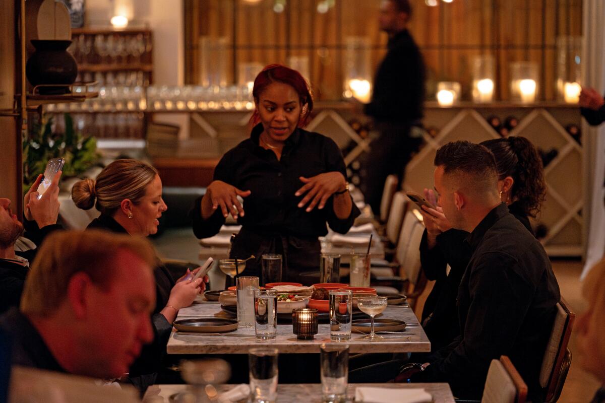 A woman in dark clothes speaks to people seated at a table in restaurant