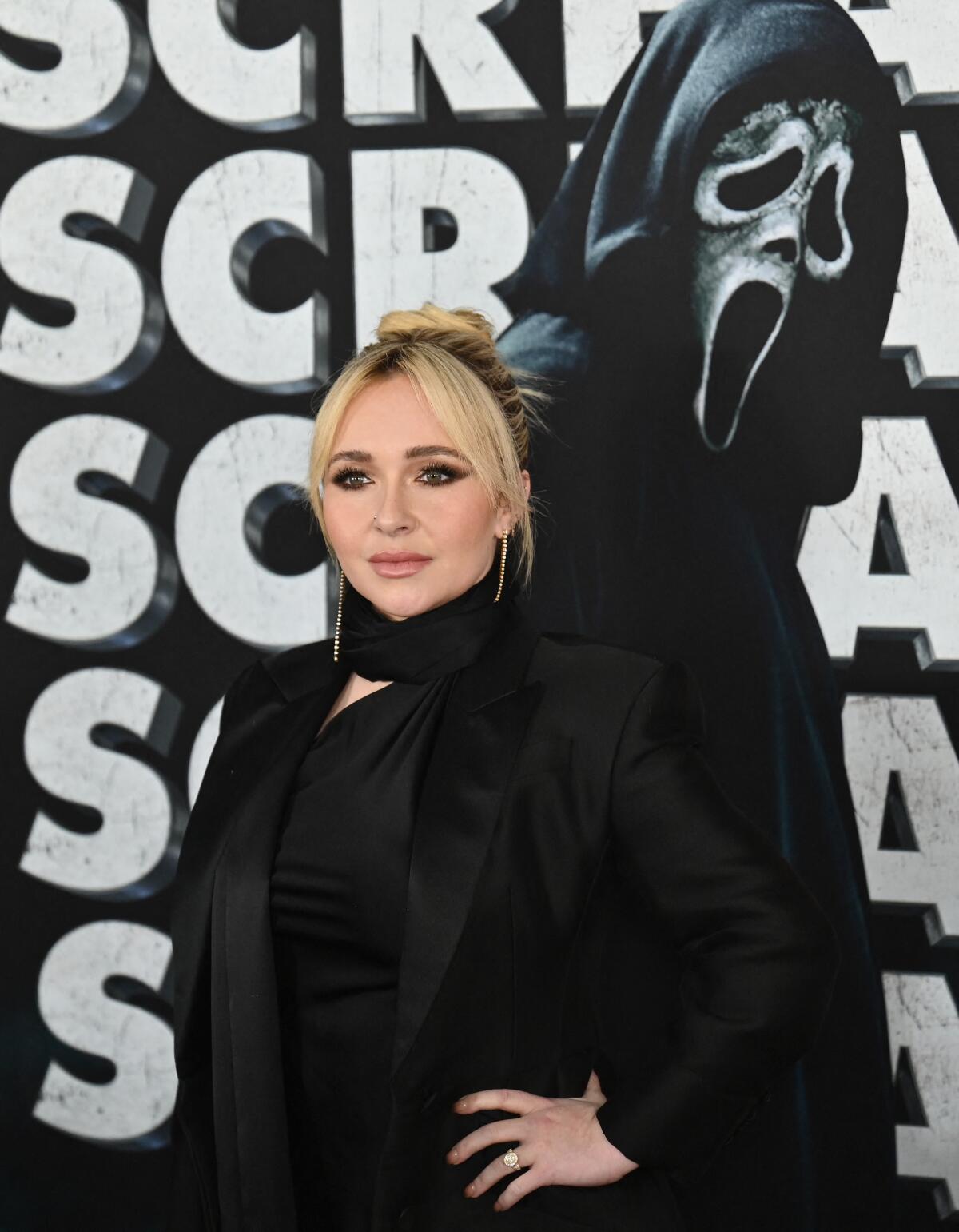 A blond woman dressed in black stands with a hand on her left hip in front of a Ghostface poster.