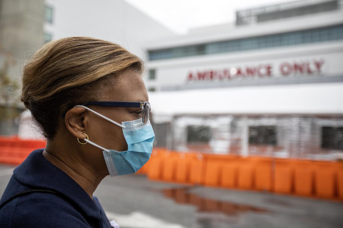 A woman wearing a mask walks around the exterior of a hospital.
