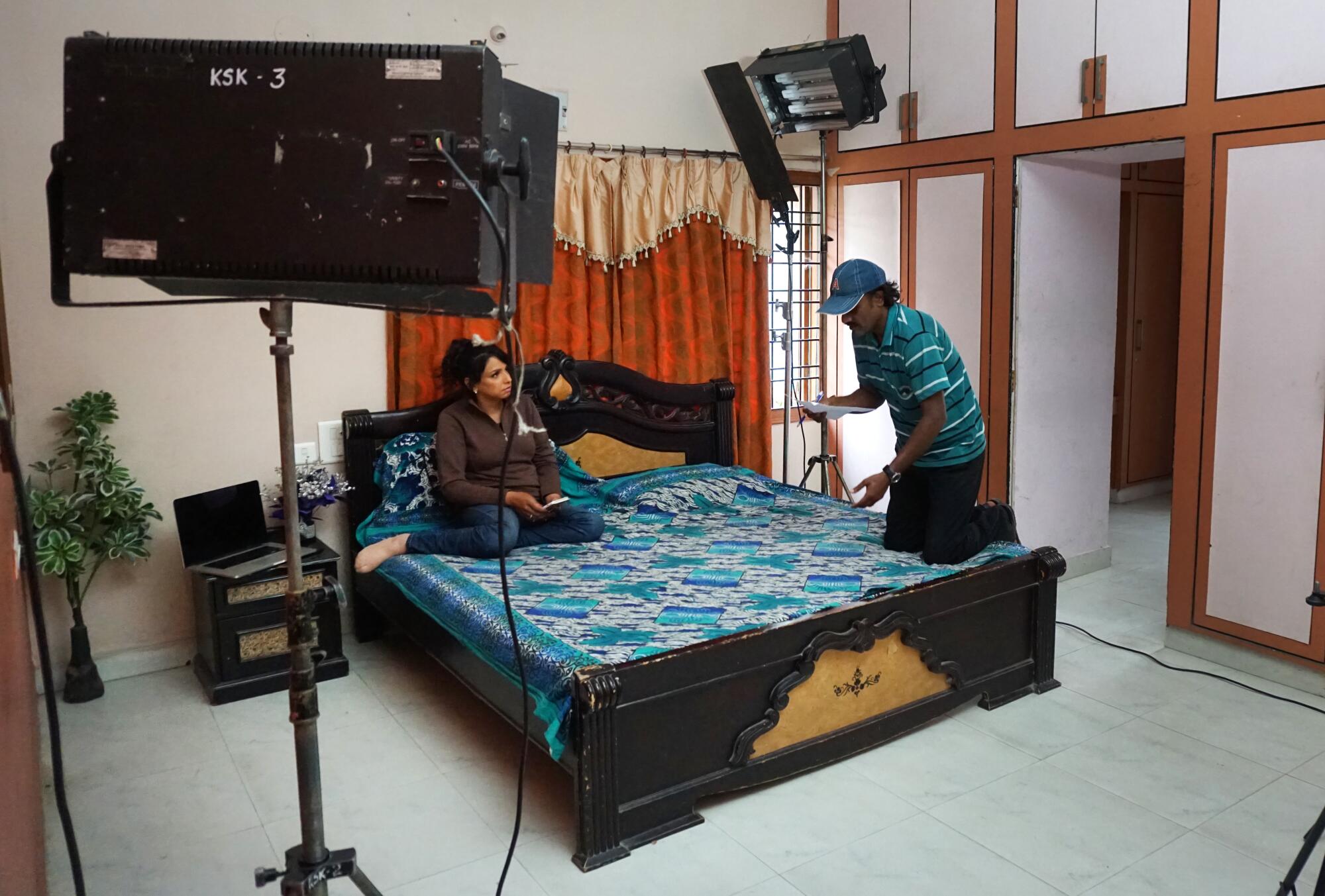 A woman sits on a bed, while a man stands nearby, with recording equipment in the room. 