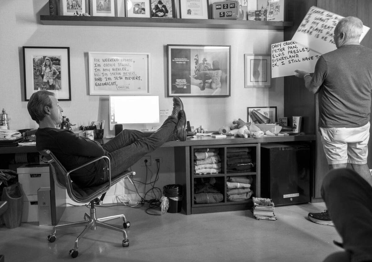 A man leans back in a desk chair while another man shows him cue cards