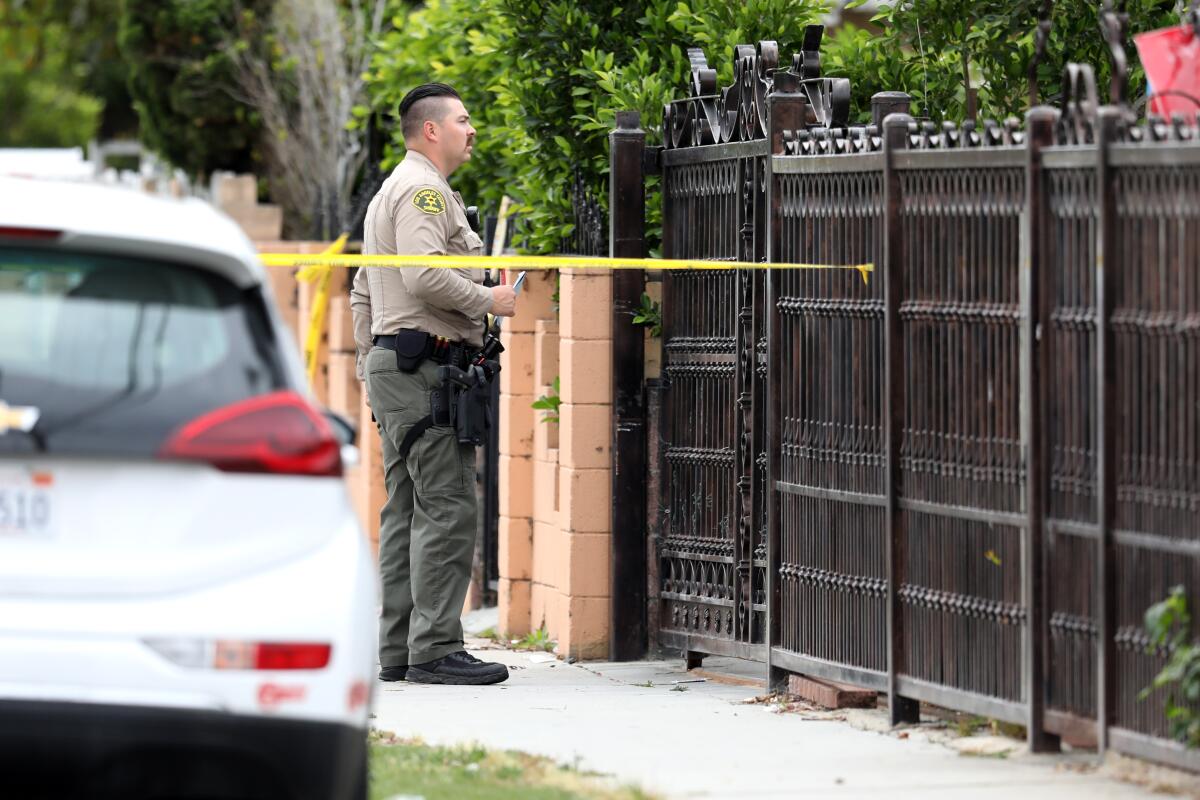 A sheriff's deputy standing behind yellow crime scene tape looks over a home's gate