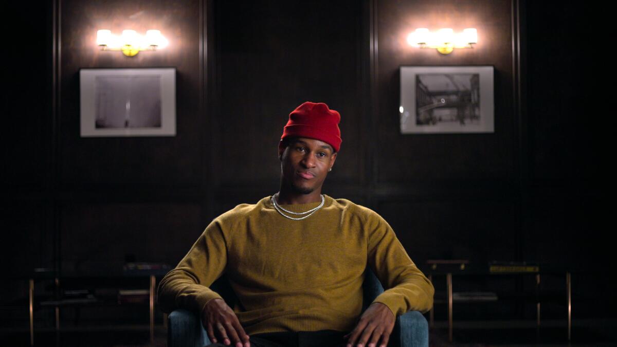 Aubrey Fisher, wearing a red beanie and olive sweater, sitting in a dark room, two framed black-and-white photos behind him.