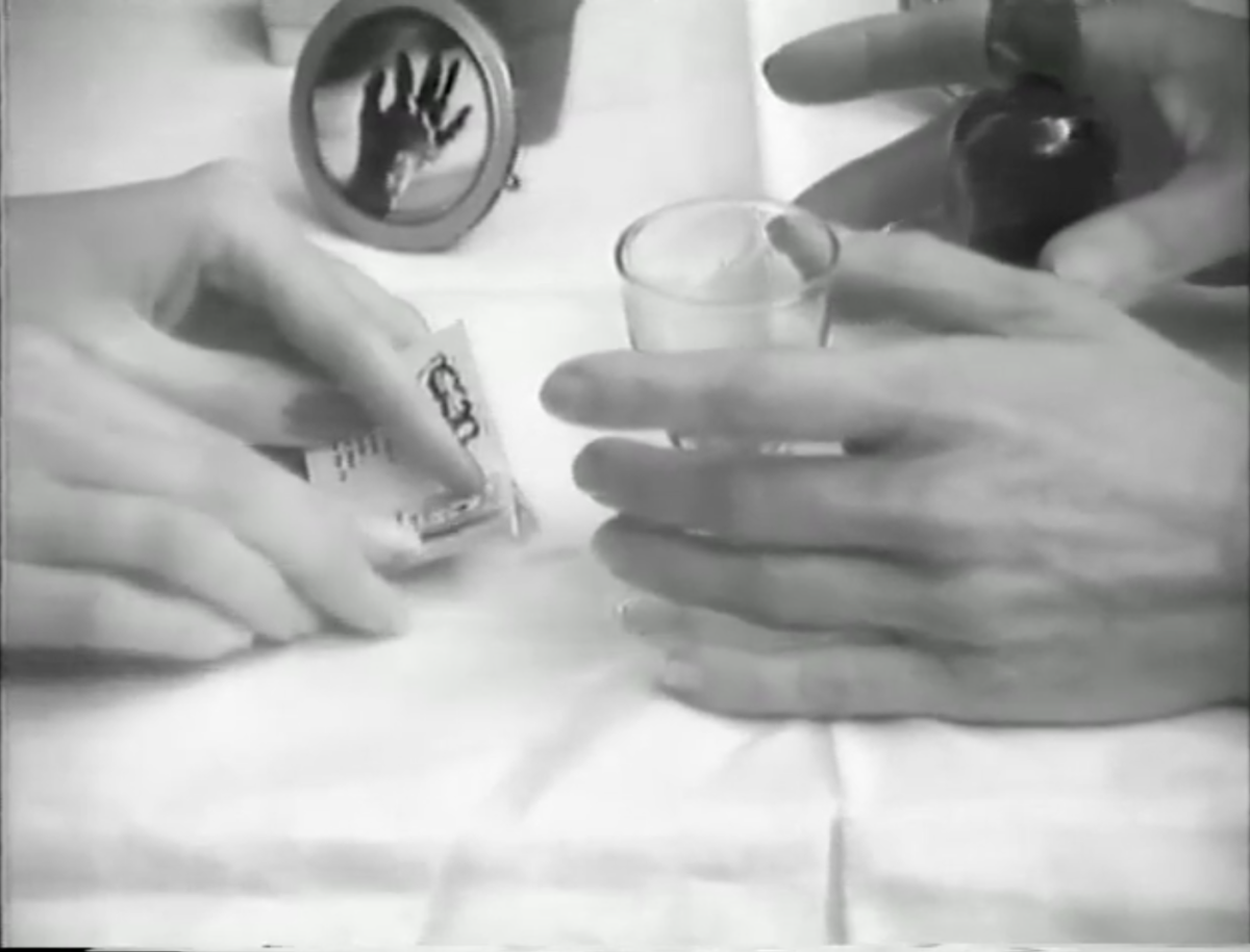 Black-and-white closeup of two pairs of hands on a white tablecloth. One is holding cards, the other a small glass.