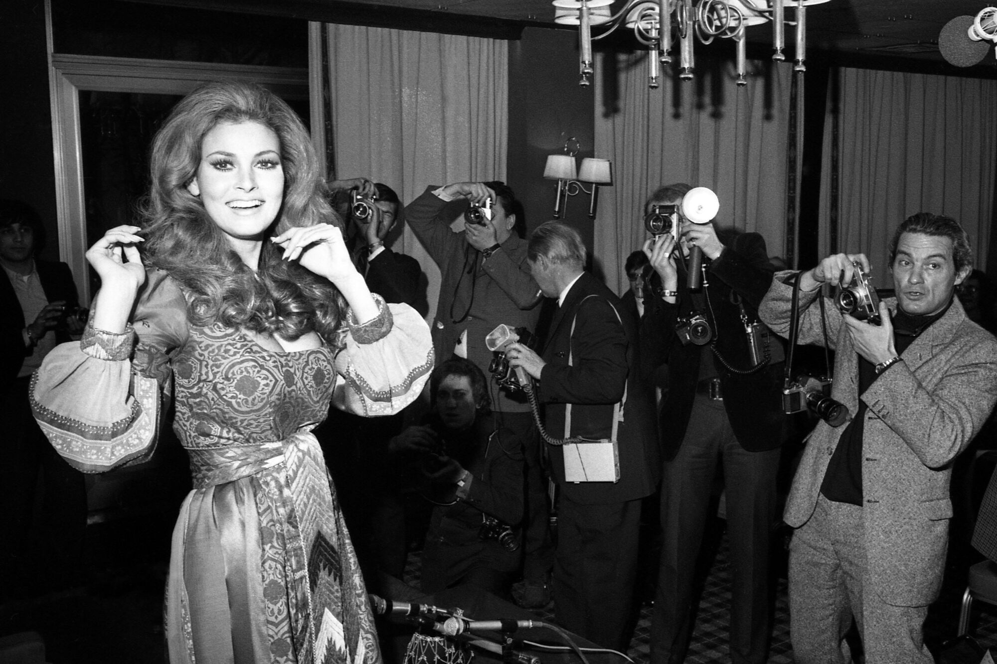 A black-and-white image of Raquel Welch, turning away from journalists to smile for the camera.
