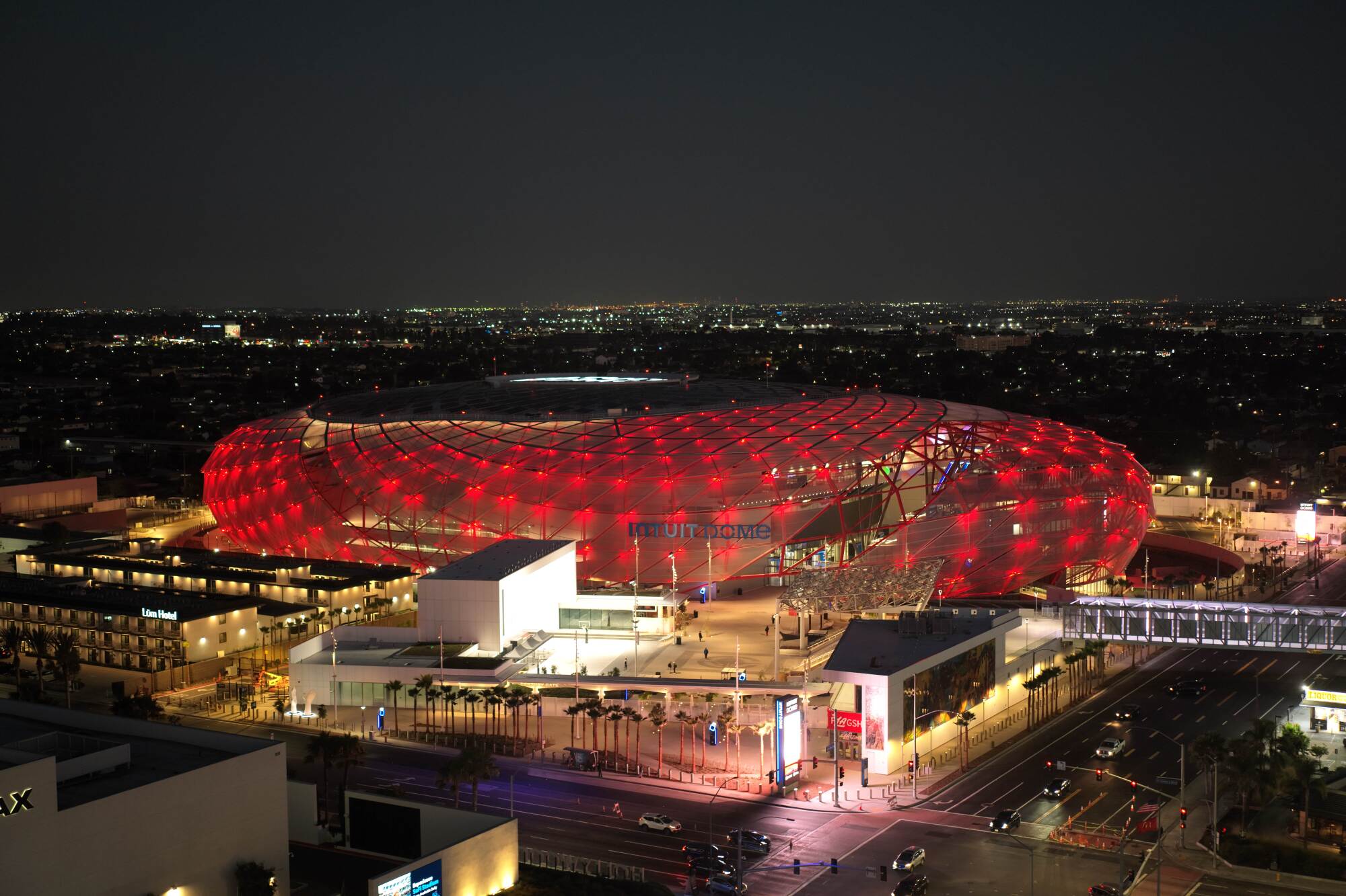 A drone image showing the Intuit Dome at night, with its translucent top glowing with red light.